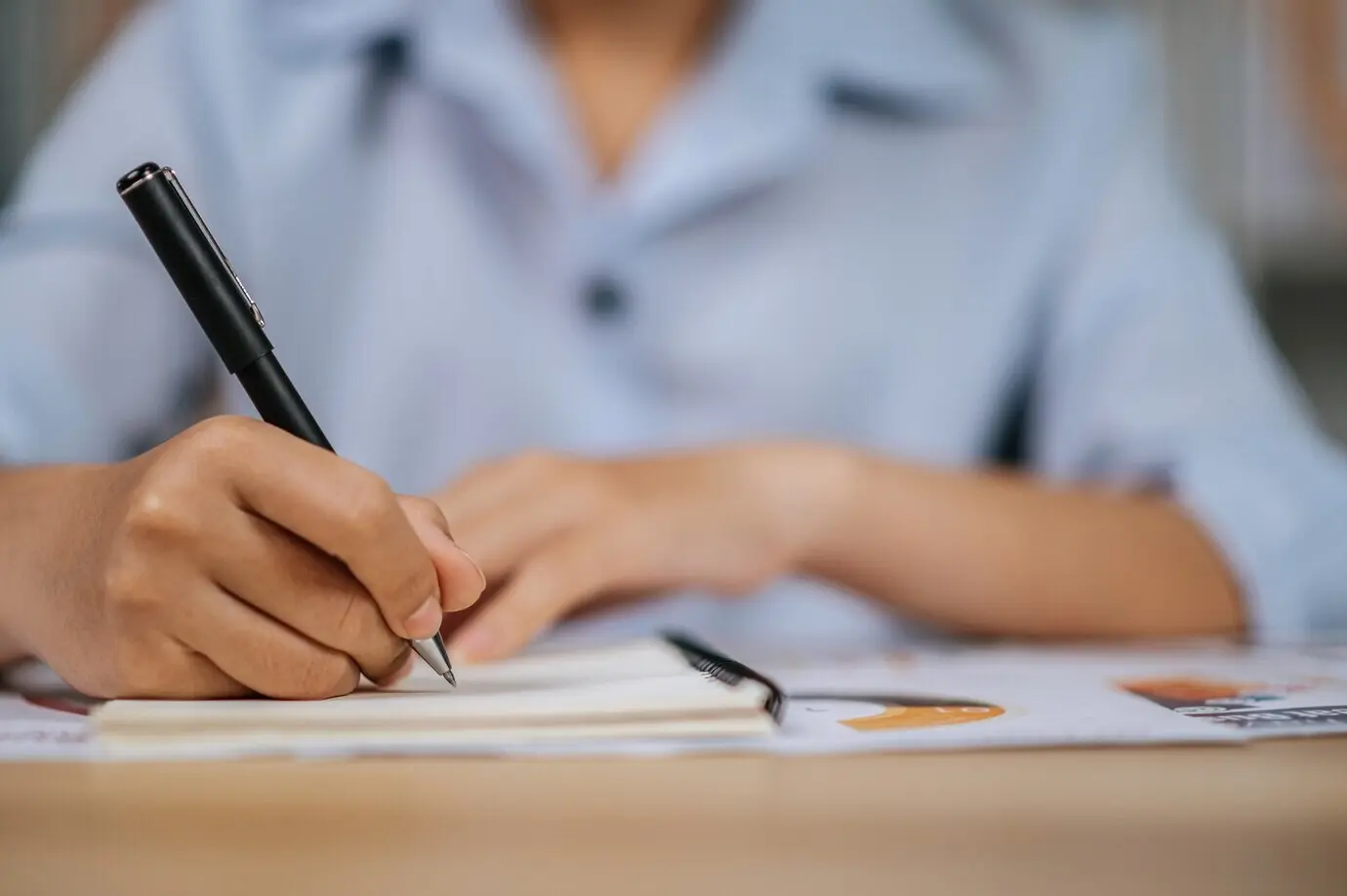 Selektiver Fokus auf die Hand einer jungen asiatischen Frau mit Brille, die mit einem Stift an Papieren im Homeoffice arbeitet, während der COVID-19-Quarantäne und Selbstisolation zu Hause; Konzept des Arbeitens von zu Hause.